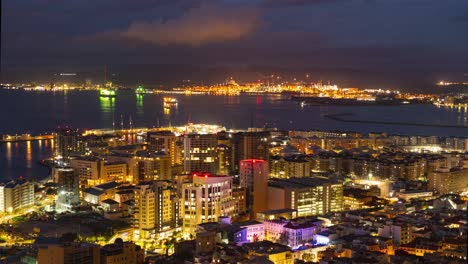 Cityscape-of-Gibraltar-and-port-behind-at-night-with-glowing-lights,-time-lapse