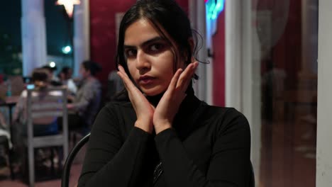 Young-woman-in-black-contemplating-at-a-cafe-during-evening,-with-dim-lights-and-customers-in-background