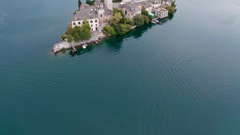 Isola-San-Giulio-En-El-Lago-Orta-En-Italia,-Mostrando-Edificios-Históricos-Y-Aguas-Tranquilas,-Colinas-Verdes-Al-Fondo,-Vista-Aérea