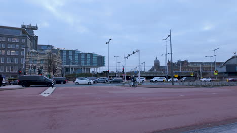 A-wide-street-with-multiple-lanes-for-cars-and-bikes,-and-a-tram-or-train-station-in-the-background