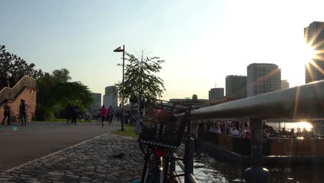 Sunset-in-Frankfurt-with-people-walking-by-the-river,-silhouette-of-a-bicycle-in-foreground,-cityscape-in-the-background