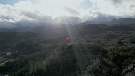 Flatternde-Flagge-Montenegros-Mit-Himmelsstrahlen,-Die-Dunkle-Wolken-Durchdringen,-Dramatisches-Luftpanorama