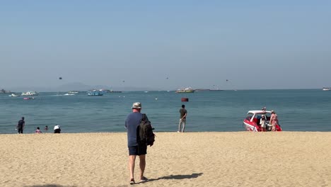 People-stroll,-watching-the-fun-of-parasailing,-and-kids-play-along-the-beach-in-Pattaya,-Thailand