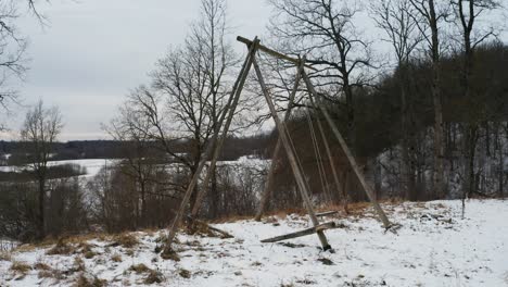 Old-wooden-swing-near-hillside-move-in-wind,-overcast-and-snowy-winter-day