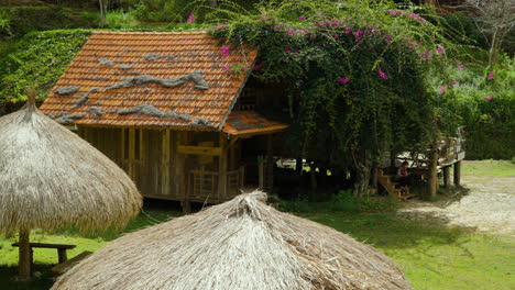 Pueblo-Folklórico-De-Cu-Lan-En-Vietnam--antiguo-Edificio-De-Madera-Con-Techo-De-Tejas-Naranjas-Y-Sombrillas-De-Paja-En-Medio-De-Una-Exuberante-Vegetación-Y-Flores