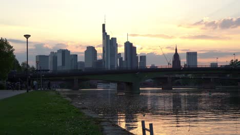 Sonnenuntergangssilhouette-Der-Frankfurter-Skyline-Mit-Reflexionen-Auf-Dem-Hauptfluss-Und-Fußgängern-Auf-Der-Brücke