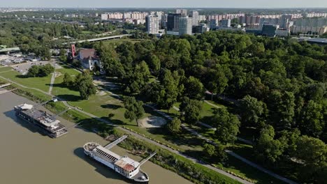 Aerial-drone-view-of-Sad-Janka-Krala---park-on-Danube-riverside-with-green-trees-and-apartment-houses-in-the-back-on-sunny-day