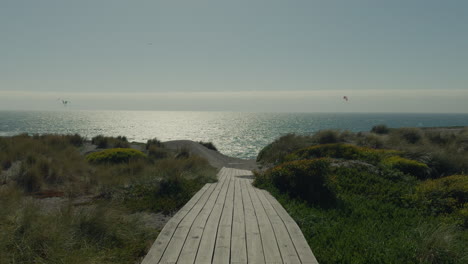 Leere-Promenade,-Die-Zum-Strand-Führt,-Mit-Ruhigem-Meer-Im-Hintergrund-Und-Der-Silhouette-Von-Kitesurfern