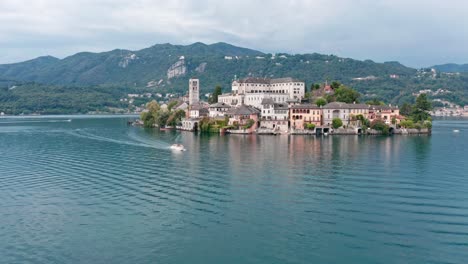 Isola-di-San-Giulio-in-Lake-Orta,-Italy,-with-historical-buildings-surrounded-by-calm-waters-and-lush-hills-in-the-background,-aerial-view