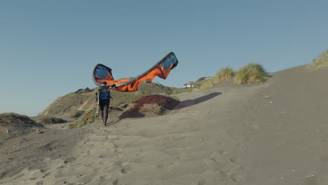 Male-Walking-Up-Beach-Path-Holding-Kite-Surf-Sail-On-Windy-Sunny-Day-In-Matanzas,-Chile