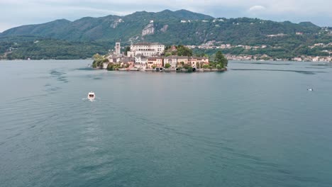 Isola-San-Giulio-on-Lake-Orta,-Italy-with-a-boat-passing-by,-green-hills-in-the-background,-aerial-view
