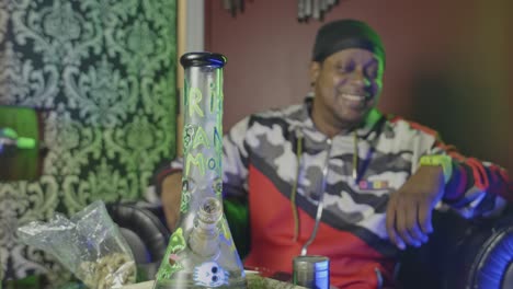 a-black-man-is-stoned-after-taking-a-hit-from-a-marijuana-ice-filled-water-bong-the-cannabis-smoking-device-is-sitting-on-the-table-with-a-bag-of-weed