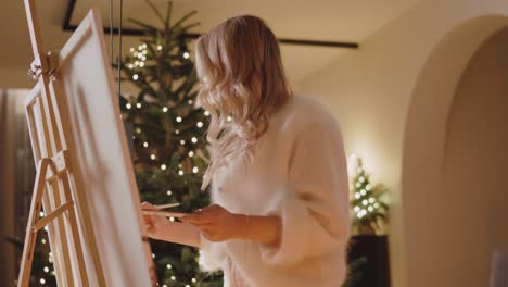 Blondes-Mädchen-Malt-Auf-Leinwand-Im-Schlafzimmer-Mit-Weihnachtsbaum-Im-Hintergrund