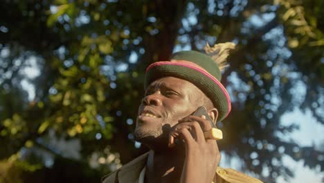 Hombre-Africano-Con-Sombrero-De-Plumas-Hablando-Por-Teléfono-Móvil-En-El-Bosque-En-Uganda