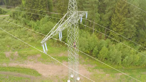 Technician-climbing-a-high-voltage-power-line-tower-amidst-a-green-forest,-aerial-view