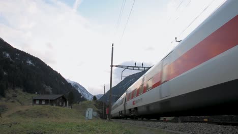 Fast-moving-train-passing-through-a-mountainous-landscape-at-dusk,-with-a-quaint-cabin-and-snowy-peaks-in-the-background