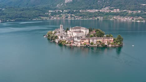 Isola-San-Giulio-on-calm-Lake-Orta,-Italy,-with-motor-boat-passing-by-and-lush-hills-in-the-background,-aerial-view