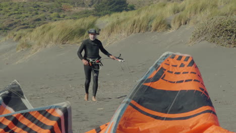 Male-In-Wetsuit-Checking-Rigging-Line-As-He-Walking-Back-To-Orange-Kitesurf-Sail-On-Windy-Beach-In-Matanzas,-Chile