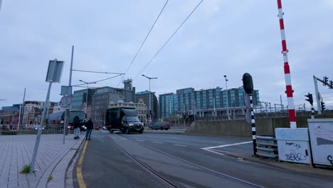 A-view-of-a-city-street-with-tram-lines,-a-railway-crossing-barrier,-and-various-urban-buildings,-possibly-near-a-main-transportation-hub-or-central-station-in-Amsterdam