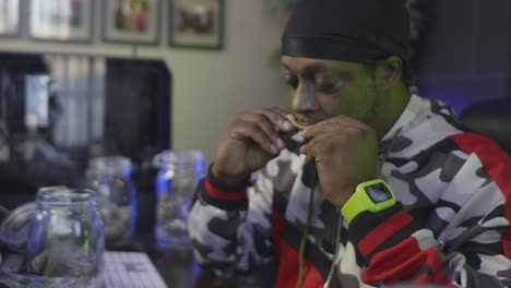 side-view-of-a-black-man-with-a-black-head-wrap-is-licking-a-blunt-to-seal-the-marijuana-into-the-tobacco-wrapper-with-mason-jars-of-cannabis-buds