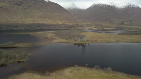 Aerial-backwards-shot-of-kilchurn-castle-at-loch-awe-lake-with-snowy-mountains-in-background,-Scotland-highlands-during-grey-sky