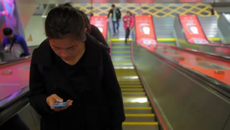 Eine-Asiatische-Frau-Blickt-Aufmerksam-Auf-Den-Bildschirm-Ihres-Telefons,-Während-Sie-Die-Rolltreppe-Hinaufsteigt