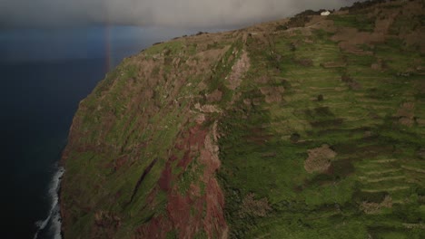 Aerial-view-with-drone-of-rugged-cliffs-beside-the-ocean-with-a-rainbow-and-waves-at-Madeira