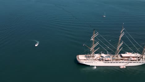 Panning-view-from-the-air-of-luxury-sailing-yacht-at-anchor-in-calm-glossy-sea-with-small-boats-and-tender-peaceful-scene-with-detail-of-decks-and-rigging