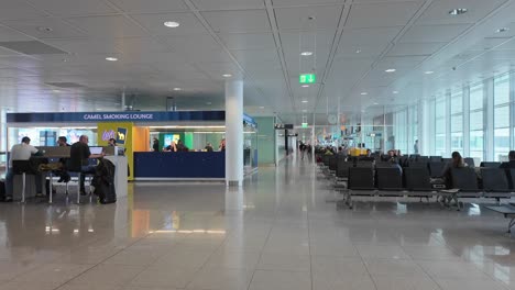 View-Inside-Munich-International-Airport-Building-In-Germany,-Passing-By-Passengers-Sitting-At-Waiting-Lounge