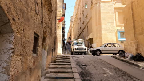 Alter-Historischer-Bürgersteig-Im-Schatten-Felsiger-Fassadengebäude,-Während-Ein-Geländewagen-Die-Straße-In-Malta-Hinunterfährt