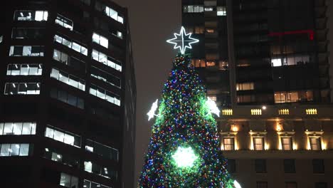 Close-up-parallax-of-Vancouver-city-very-large-and-tall-christmas-tree-lit-up-and-glowing-focused-on-the-star-on-top-surrounded-by-the-tall-lit-up-buildings-on-a-winter-night-in-the-dark