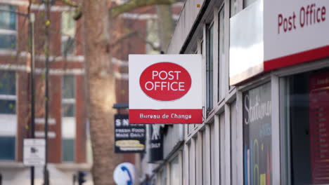 A-red-bus-passes-in-the-background-of-a-Post-Office-sign,-a-public-facility-that-provides-mail-and-other-services