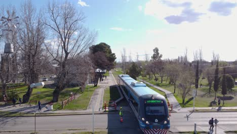 Modern-tram-crossing-an-urban-park-area-with-people-and-trees,-in-the-province-of-Neuquén,-Argentina