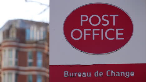 The-corner-of-a-red-brick-ornate-building-stands-behind-a-red-and-white-Post-Office-and-Bureau-de-Change-logo-on-a-sign,-a-public-facility-that-provides-mail-and-other-services