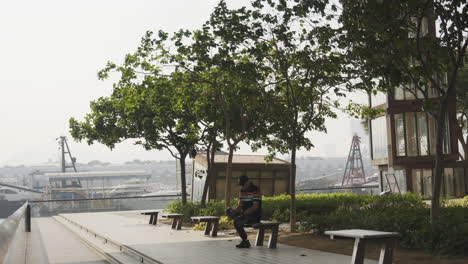 Old-asian-man-sits-on-modern-concrete-park-bench-overlooking-city-in-shade-of-trees-on-windy-day