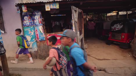 Vista-En-Cámara-Lenta-De-Niños-Caminando-Con-Mochilas-Y-Una-Pareja-Cortando-Leña-Afuera-De-Una-Pequeña-Tienda-En-Una-Calle-Típica-En-El-Pueblo-De-Guatemala