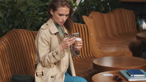 Kaukasische-Studentin-Benutzt-Smartphone-Und-Trinkt-Kaffee-Im-Freien.