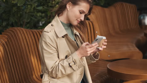 Caucasian-female-student-using-smartphone-and-drinking-coffee-outdoors.