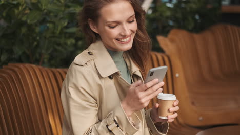 Caucasian-female-student-using-smartphone-and-drinking-coffee-outdoors.