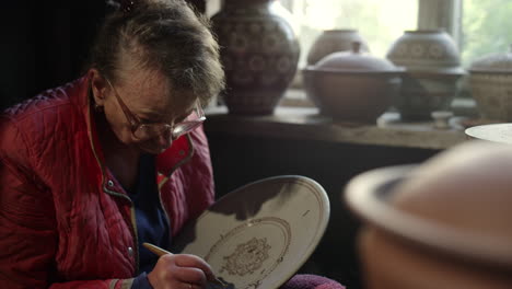 Serious-old-woman-decorating-plate-in-pottery