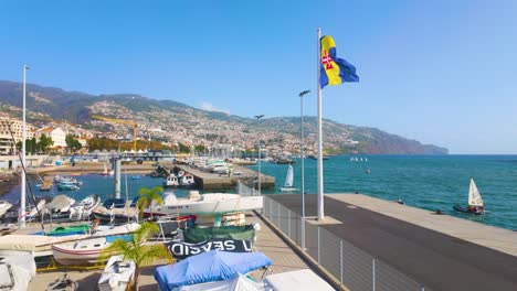 Waving-flag-of-Funchal-at-harbor-with-docking-boats-and-mountains-in-Background-during-sunny-day---Portugal,Europe