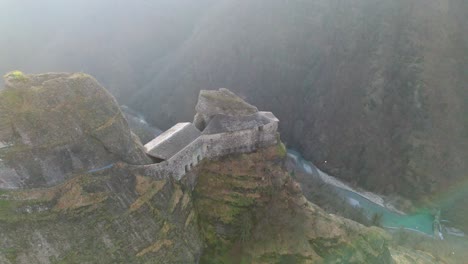 Majestic-ancient-fortress-atop-a-rugged-cliff-with-a-river-below-in-the-misty-morning-light,-aerial-view