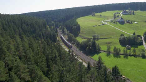 Aerial-view-of-a-red-Deutsche-Bahn-train-traversing-lush-green-landscape-with-dense-forests-and-rural-homes-in-daylight