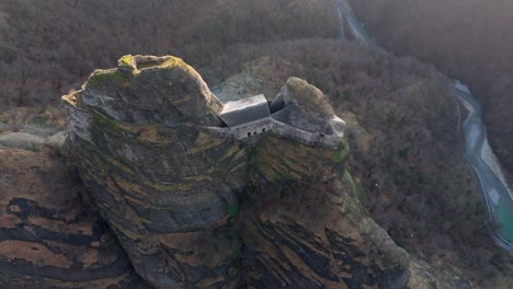Ancient-castle-atop-a-rugged-cliff-with-a-river-winding-in-the-background-at-dusk,-aerial-view