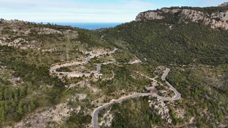Aerial-view-showing-serpentine-road-on-green-hill-of-Esporles-on-Mallorca-and-ocean-in-background