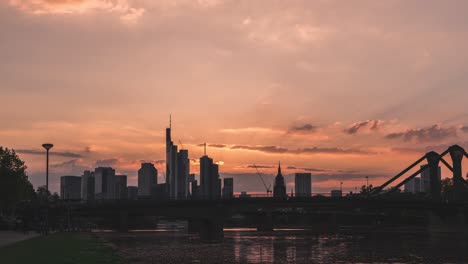 Silhouette-of-Frankfurt-skyline-against-a-sunset-sky-with-clouds-and-bridge-over-river,-timelapse