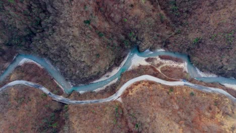 Winding-road-tracing-through-a-rugged-autumn-landscape-parallel-to-a-river,-top-down-aerial-view