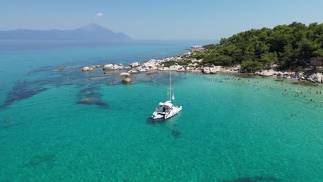 Barco-Catamarán-Y-Gente-Nadando-En-La-Bahía-Azul-Turquesa-En-Halkidiki,-Grecia---Círculos-Aéreos-4k