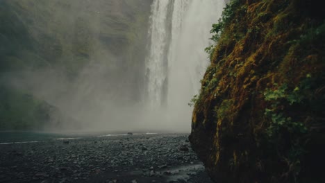 Reveal-shot-from-behind-cliffs-of-scenic-misty-Skógafoss-falls-in-Iceland,-slomo