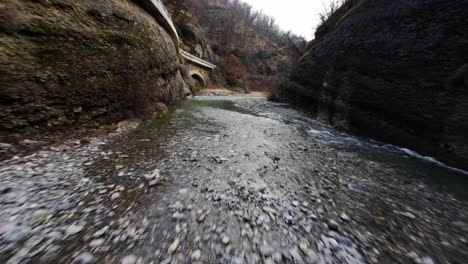 Rushing-river-through-a-narrow-rocky-canyon-with-pebbled-shore,-captured-in-natural-daylight,-FPV-shot